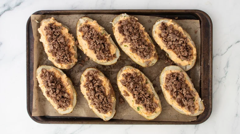 Potato skins on a large baking sheet filled with seasoned mashed potato and taco beef