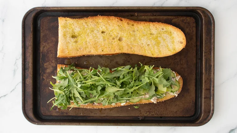 A layered tuna salad on a large toasted baguette topped with baby arugula.